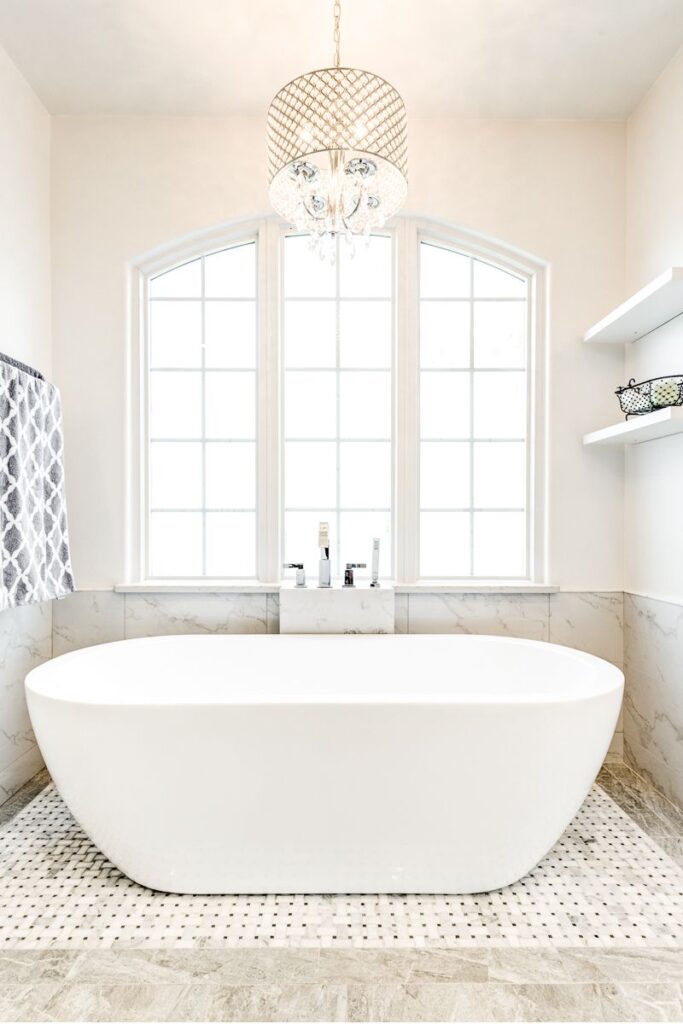 Modern bathroom with a freestanding white bathtub, marble tile flooring, large windows, and a decorative chandelier.