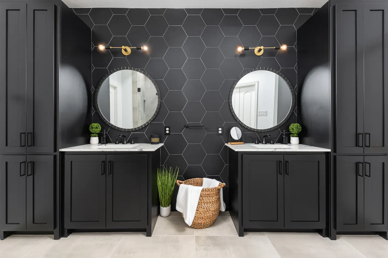 Black double vanity with round mirrors and brass sconces on a black hexagon tile wall, between tall black cabinets