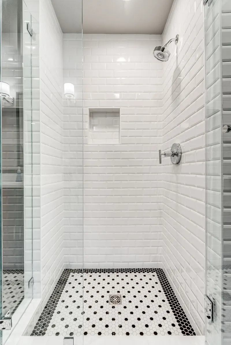 Prosper walk-in shower by Nomi with white beveled subway tile, frameless glass and a black and white mosaic floor